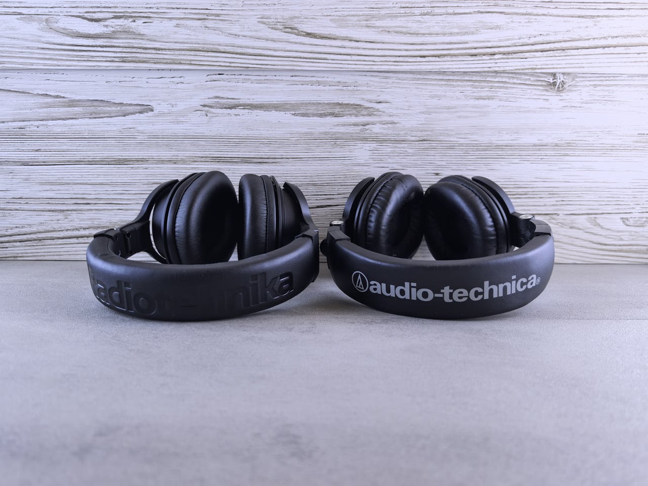 Two black Audio-Technica headphones are placed side by side on a light grey surface with a wooden background, highlighting the brand’s logo—a stylish reminder to protect your ears and watch for early signs of hearing loss in adults.